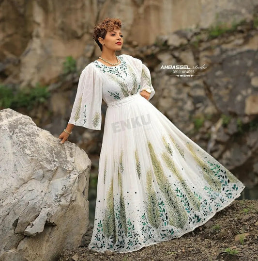A woman wearing a flowing white maxi Green Habesha Dress  with green and beige floral embroidery and beadwork, standing outdoors against a rocky cliff background. The dress has three-quarter sleeves and a fitted waist.