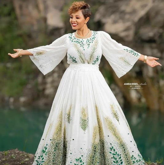A woman wearing a long white maxi Green Habesha Dress  with green and gold leaf patterns, standing outdoors with her arms outstretched. The background shows a blurred natural setting with green water and brown rocks.