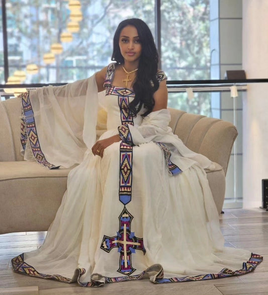 A woman sits on a beige sofa wearing a flowing white traditional Ethiopian Dress with colorful geometric embroidered details and a large cross motif on the front. The dress has sheer draped sleeves and a long train spread across a light floor in a modern indoor setting.
