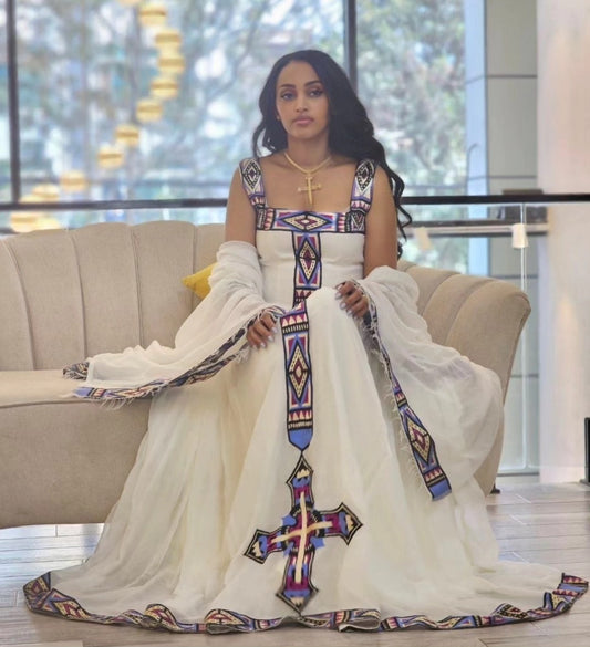 A woman seated on a beige sofa wearing a voluminous white Ethiopian Dress  with colorful geometric embroidery and a large cross motif at the center front. The dress has sheer draped sleeves and a long train, photographed in a bright indoor setting with large windows behind her.