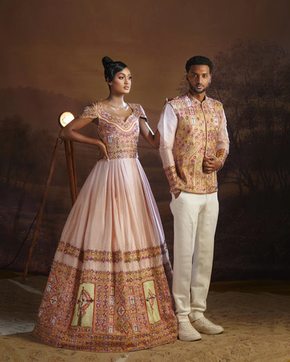 A man and a woman pose in traditional Ethiopian Wedding matching couple set. The woman wears a light pink Anarkali gown with intricate gold and multicolor embroidery, and the man wears a matching embroidered vest over a white shirt and white trousers.