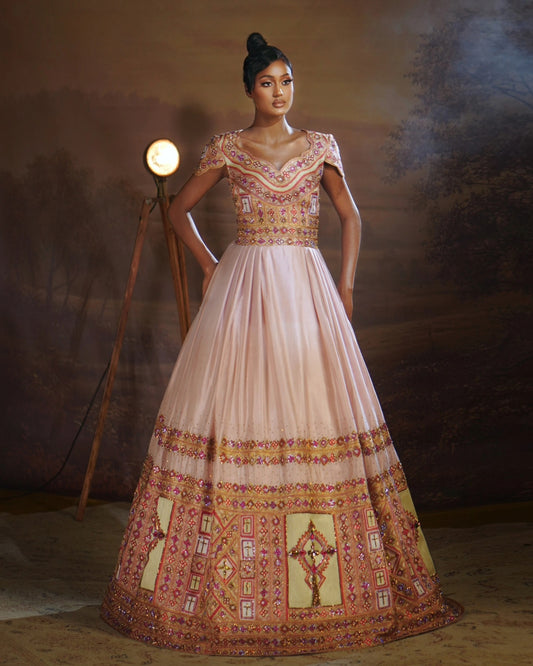 A woman modeling a traditional Indian Ethiopian Wedding matching couple set with a light pink skirt and heavily embroidered bodice and hem in gold, red, and green, set against a dark, moody studio backdrop with a vintage floor lamp.