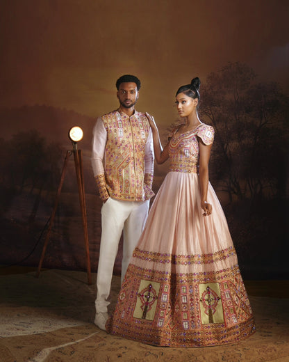 A man and a woman pose in traditional Ethiopian Wedding matching couple set The woman wears a peach-pink lehenga choli with intricate gold, red, and green embroidery, paired with a matching pleated skirt.