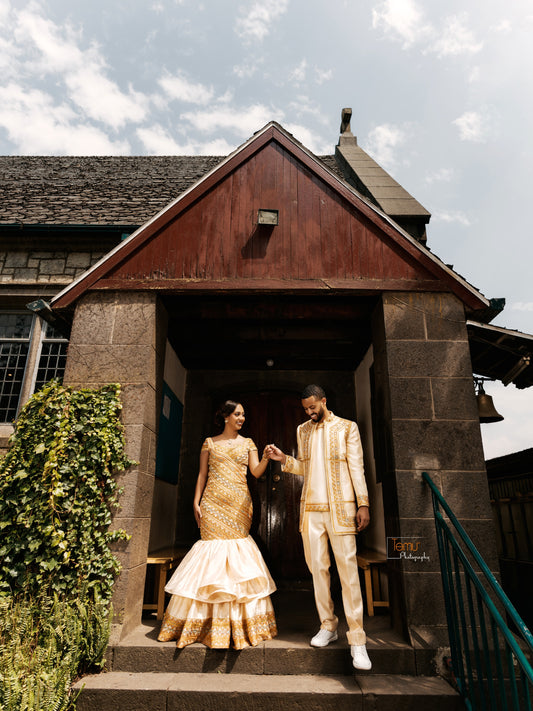 A couple posing in traditional African Ethiopian matching couple set outside a stone building with a wooden gable. The woman wears a gold and white tiered gown, and the man wears a cream and gold embroidered suit.