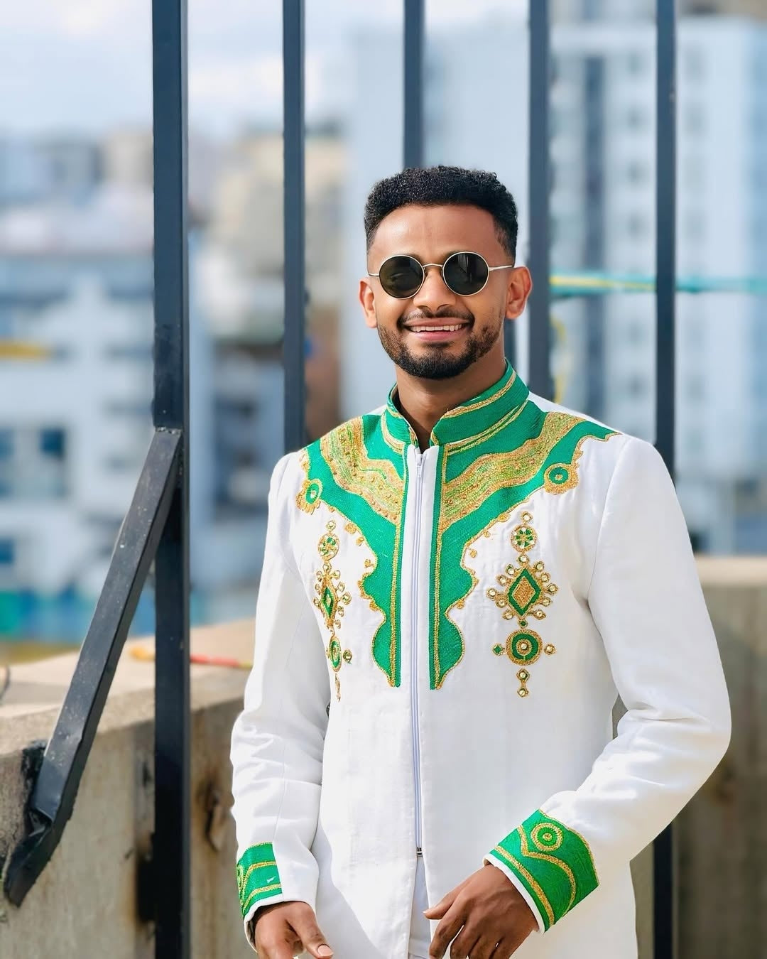 Man wearing a white and green Ethiopian mens shirt with sunglasses, standing against a blurred urban background.