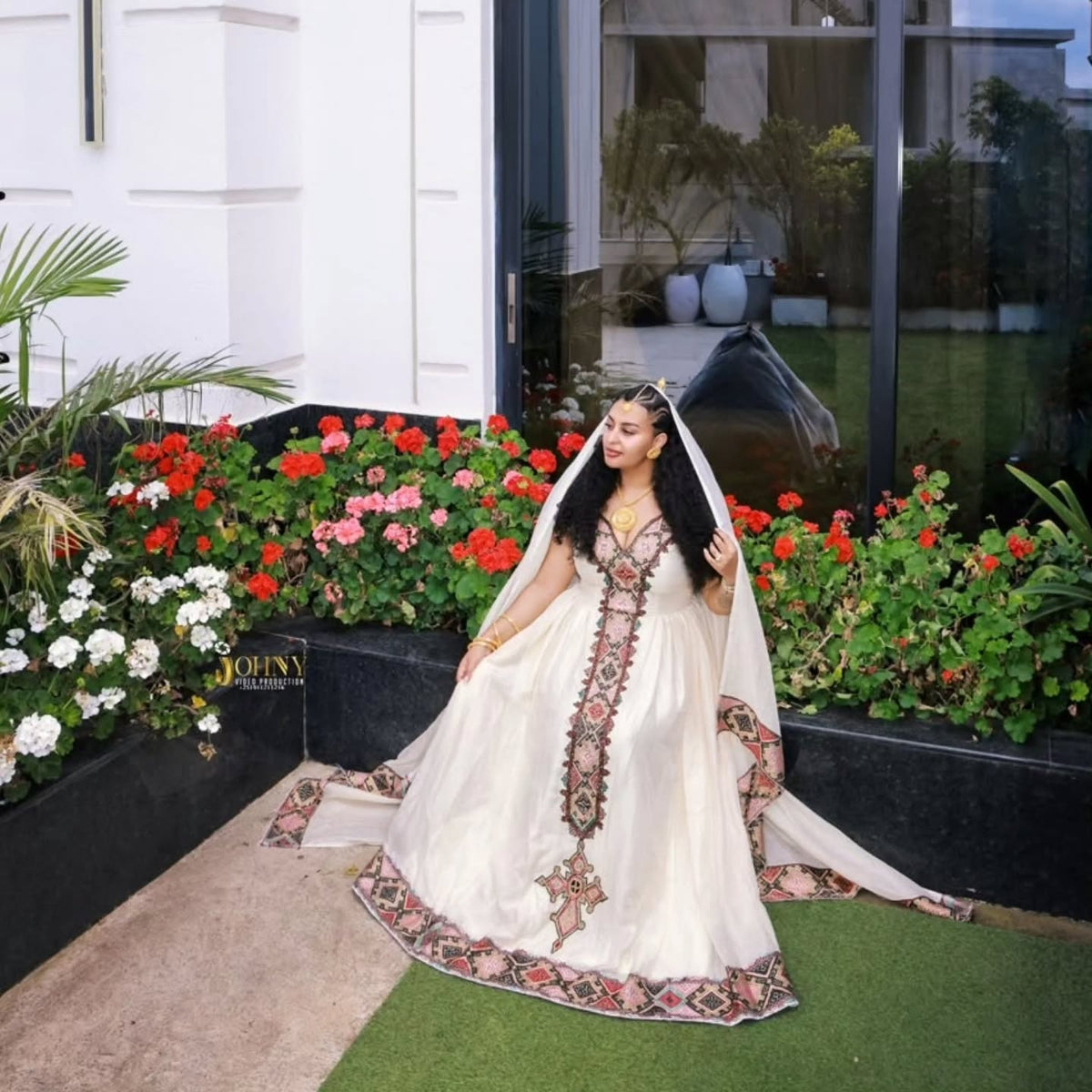 A woman wearing a traditional white embroidered Ethiopian Dress with a matching sheer veil, standing outdoors in front of a modern building with large windows and flower beds filled with red, pink, and white flowers.