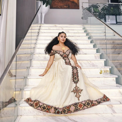 A woman wearing a voluminous off-the-shoulder cream-colored Ethiopian Dress with intricate multicolor geometric embroidery and a matching embroidered sash, posing on a modern marble staircase with glass railings.