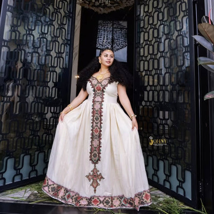 A woman models a traditional Moroccan-style white Ethiopian Dress with intricate multicolor embroidery and a large puff skirt, standing in front of a black metal gate.