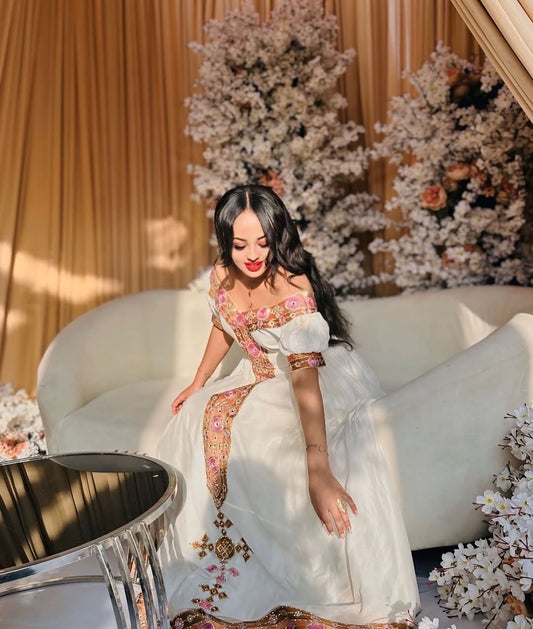 Woman in a white and gold Ethiopian Habesha Dress sitting on a white couch with floral decorations in the background.