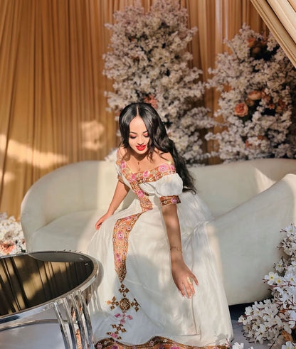 Woman in a white and gold Ethiopian Habesha Dress sitting on a white couch with floral decorations in the background.