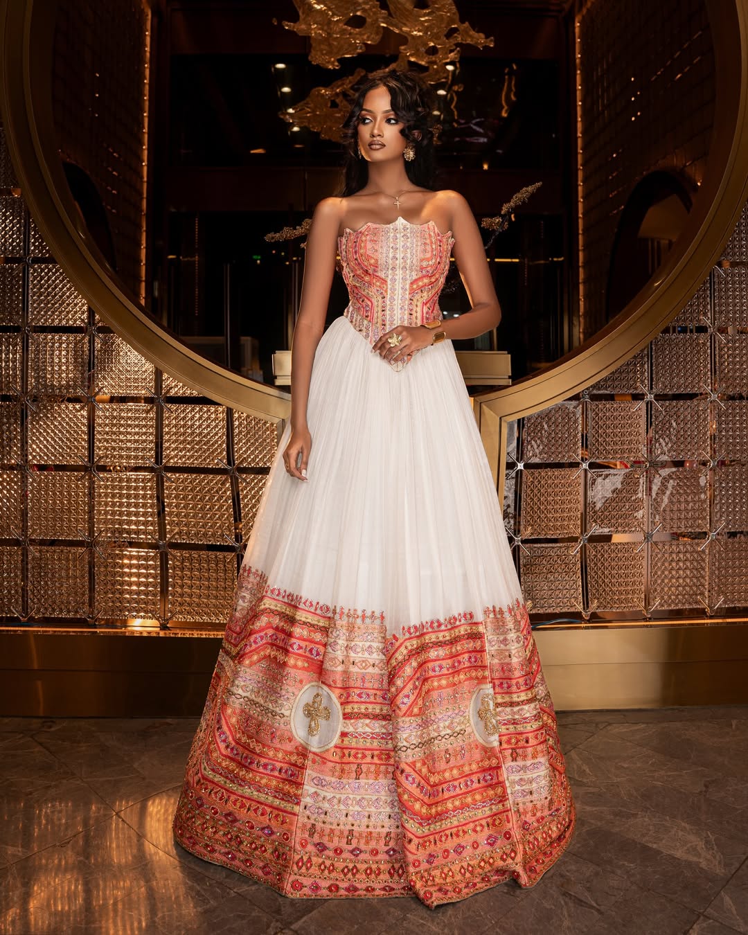 Woman wearing a traditional Ethiopian traditional dress  with a patterned top and white skirt in an elegant interior setting.