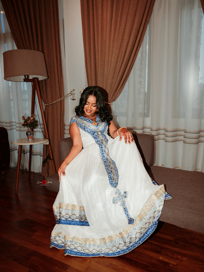 A woman wearing a white traditional Ethiopian Dress with blue and beige embroidered borders, posing in a room with brown and white curtains, a floor lamp, and a sofa.