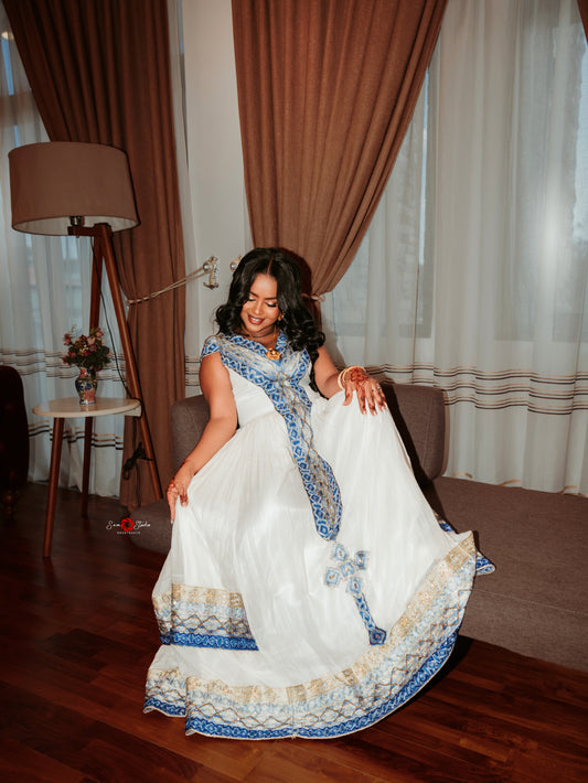 A woman wearing a white traditional Ethiopian Dress with blue and beige embroidered borders, posing in a room with brown and white curtains, a floor lamp, and a sofa.