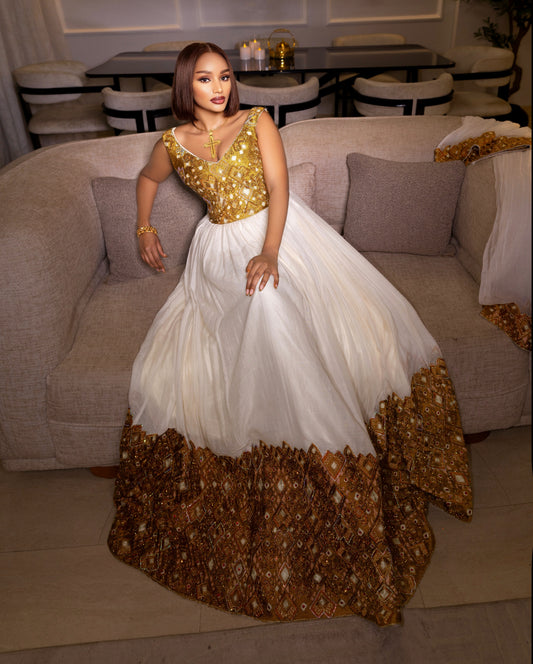 A woman wearing a traditional African Habesha Kemis  with a gold embroidered bodice and a white skirt with a gold patterned hem, sitting on a beige sofa in a living room.