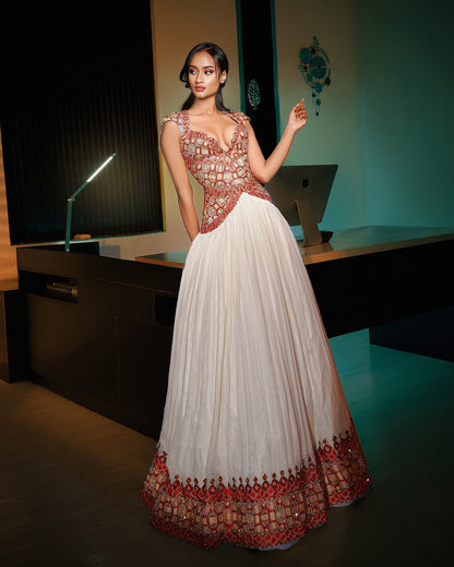 A model wears a floor-length white Ethiopian National Dresses with a heavily embellished red and gold beaded bodice and a matching embroidered hem, standing in a dimly lit modern room with a desk and computer in the background.
