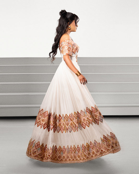 A woman modeling a floor-length off-the-shoulder Ethiopian Traditional Clothing with a cream base and extensive rust red, orange, and gold geometric embroidery concentrated on the bodice and hem, shown against a minimalist gray studio background.