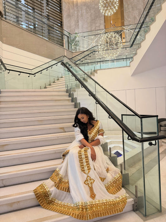 A woman wearing a white traditional Habesha Store  with elaborate gold embroidery sits on the steps of a modern glass-rail staircase in a high-rise building.