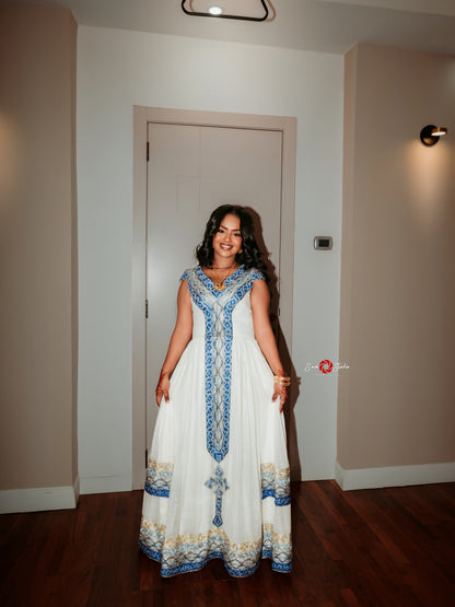 A woman stands indoors wearing a long white traditional Ethiopian Dress with blue and gold embroidered detailing. The photo is taken in a hallway with beige walls, a closed door behind her, and dark wood flooring.