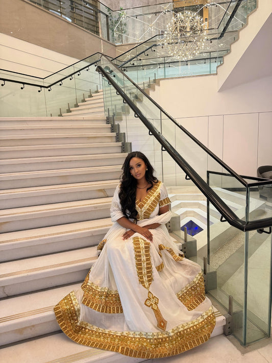 A woman wearing a white traditional Habesha Store  with ornate gold embroidery sits on a grand indoor staircase with glass railings and a modern chandelier in the background.