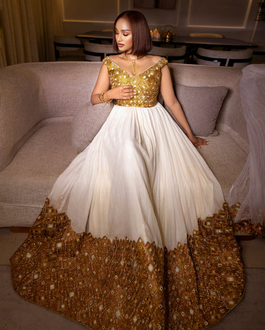 A woman wearing a traditional Indian Anarkali Habesha Kemis  with a gold embellished bodice and a voluminous off-white skirt featuring a heavily embroidered gold border, posed on a beige sofa.