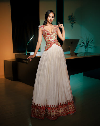 A woman models a floor-length white Ethiopian National Dresses with ornate red and gold embroidery, standing in a modern interior setting with a desk and computer in the background.