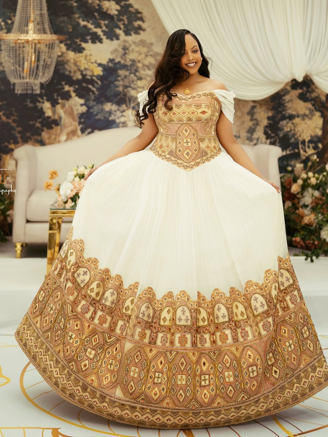 Woman in a white and gold embroidered Gold Ethiopian Wedding Dress in an elegant room with chandelier and floral decorations.
