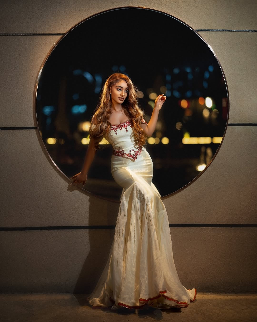 Woman in a white Red Eritrean Dress with red details sitting in front of a round mirror.