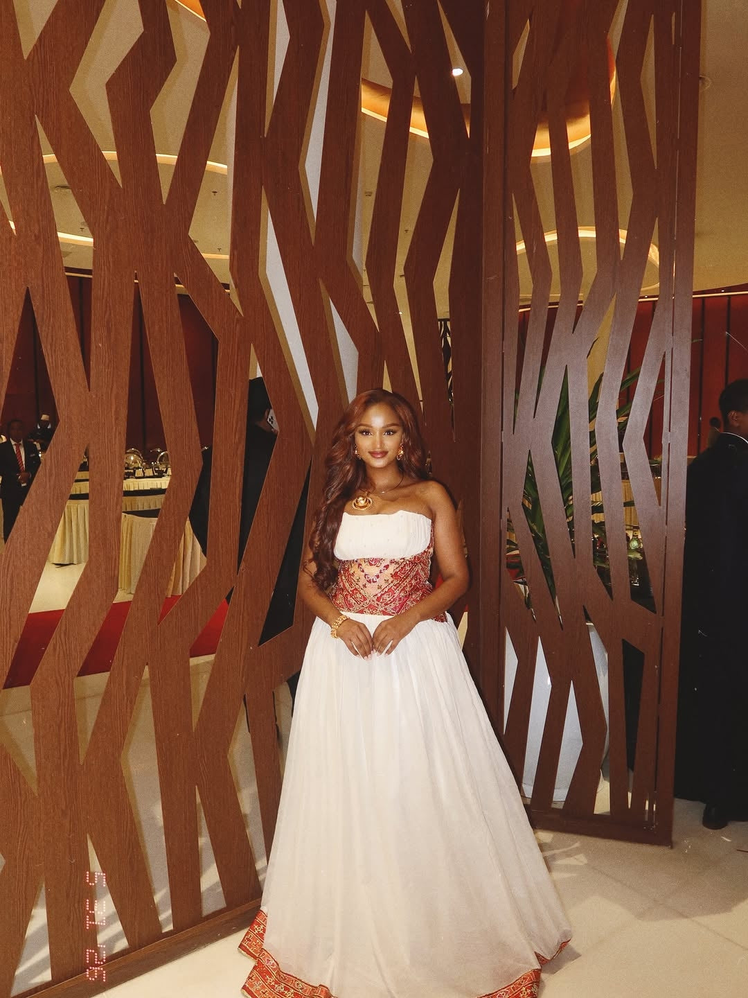 Woman in a white and red Ethiopian Traditional Dress standing in front of a decorative wooden screen.