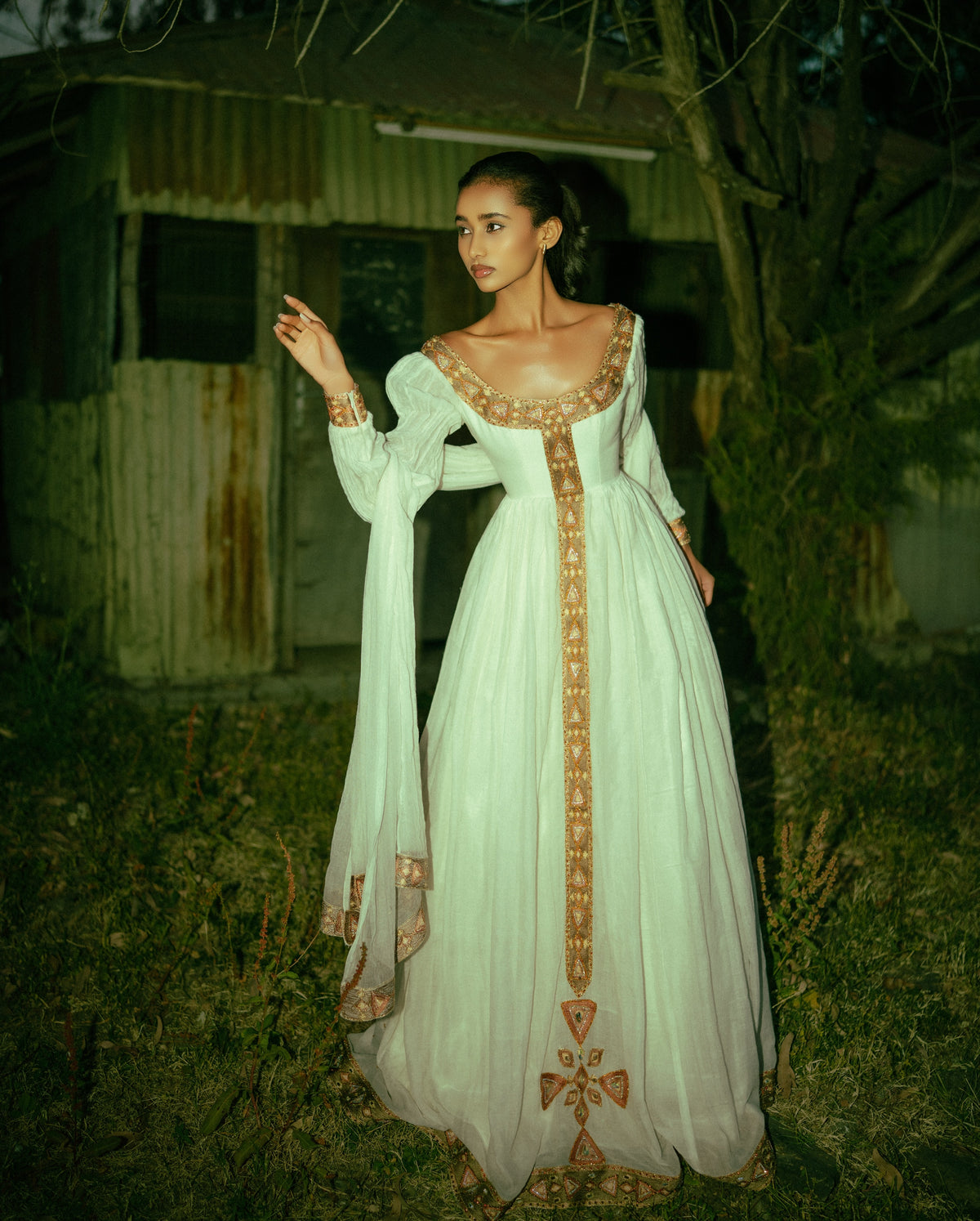 A woman stands outdoors at night wearing a long white or off-white  Habesha traditional dress  with ornate gold embroidery and a deep neckline. The dress has long sheer sleeves and a full skirt, with gold detailing running down the front and around the neckline.
