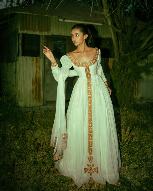 A woman stands outdoors at night wearing a long white or off-white  Habesha traditional dress  with ornate gold embroidery and a deep neckline. The dress has long sheer sleeves and a full skirt, with gold detailing running down the front and around the neckline.
