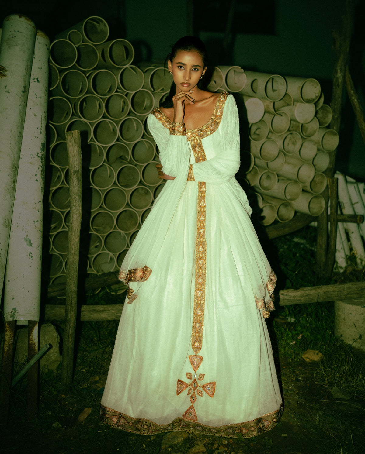 A woman wearing a long white  Habesha traditional dress with ornate gold embroidery and a deep neckline, standing outdoors in front of stacked pipes at night.