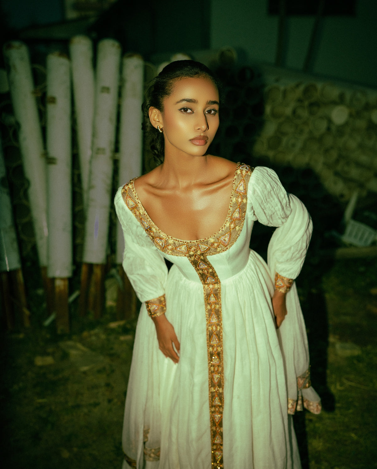 A woman wearing a long  Habesha traditional dress  with ornate gold embroidery and a deep scoop neckline, standing outdoors at night with a rustic background.