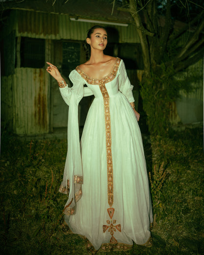 A woman wearing a long white  Habesha traditional dress with ornate gold embroidery and a sheer draped overlay, standing outdoors at night in front of a rustic building and trees.