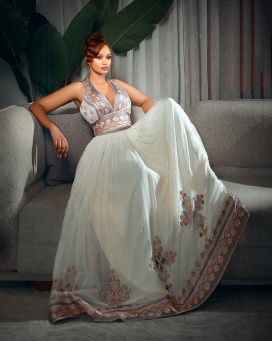 A woman models a floor-length white Ethiopian Traditional Clothing with metallic gold and silver embroidery, seated on a gray sofa in a styled indoor setting with a large green plant in the background.