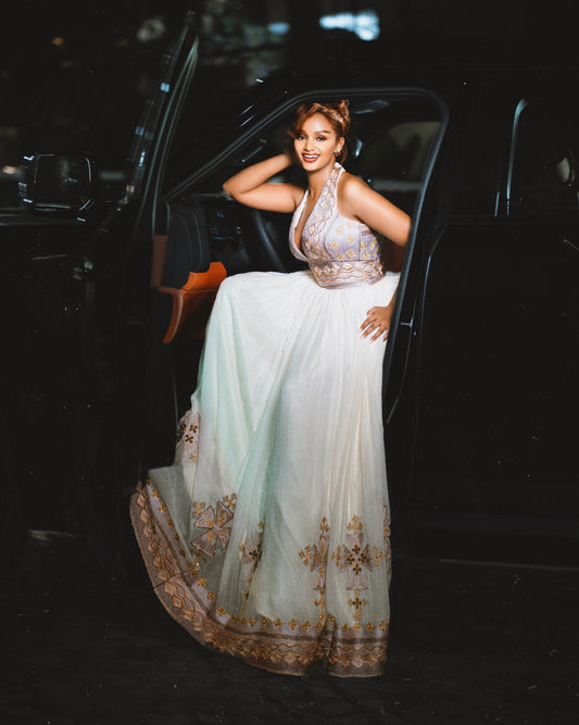 A woman posing in a white traditional Ethiopian Traditional Clothing with gold and silver embroidery, leaning against the open door of a black car at night.