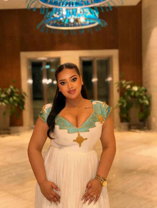 Woman in a white and Green Ethiopian Dress with gold accents standing in a decorated indoor setting.