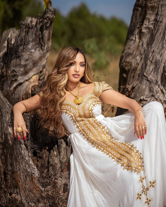 Woman in a white and gold habesha libs sitting between two trees outdoors.