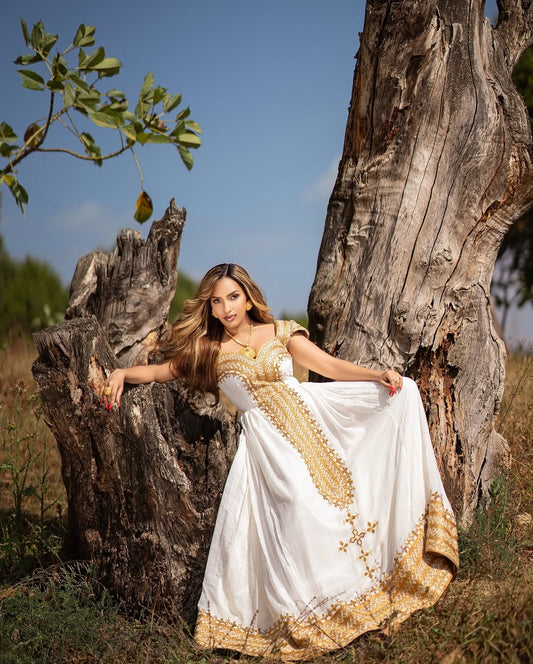 Woman in a white and gold habesha libs sitting between two large trees in a natural setting.
