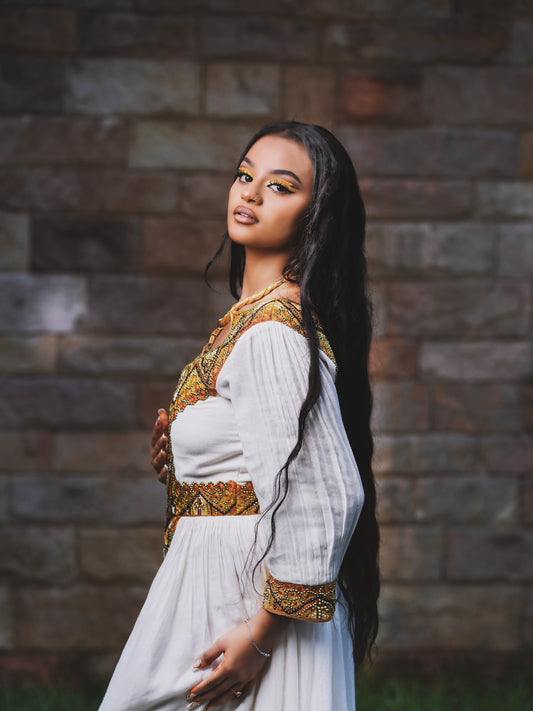 A woman with long dark hair wearing a white Ethiopia clothes with ornate gold and brown embroidery, standing in front of a rustic brick wall.