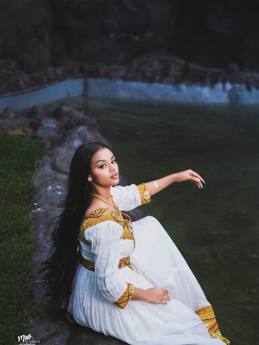 A woman with long black hair sits by a body of water wearing a white Ethiopia clothes with ornate gold embroidery. The background features dark rocks and green grass.