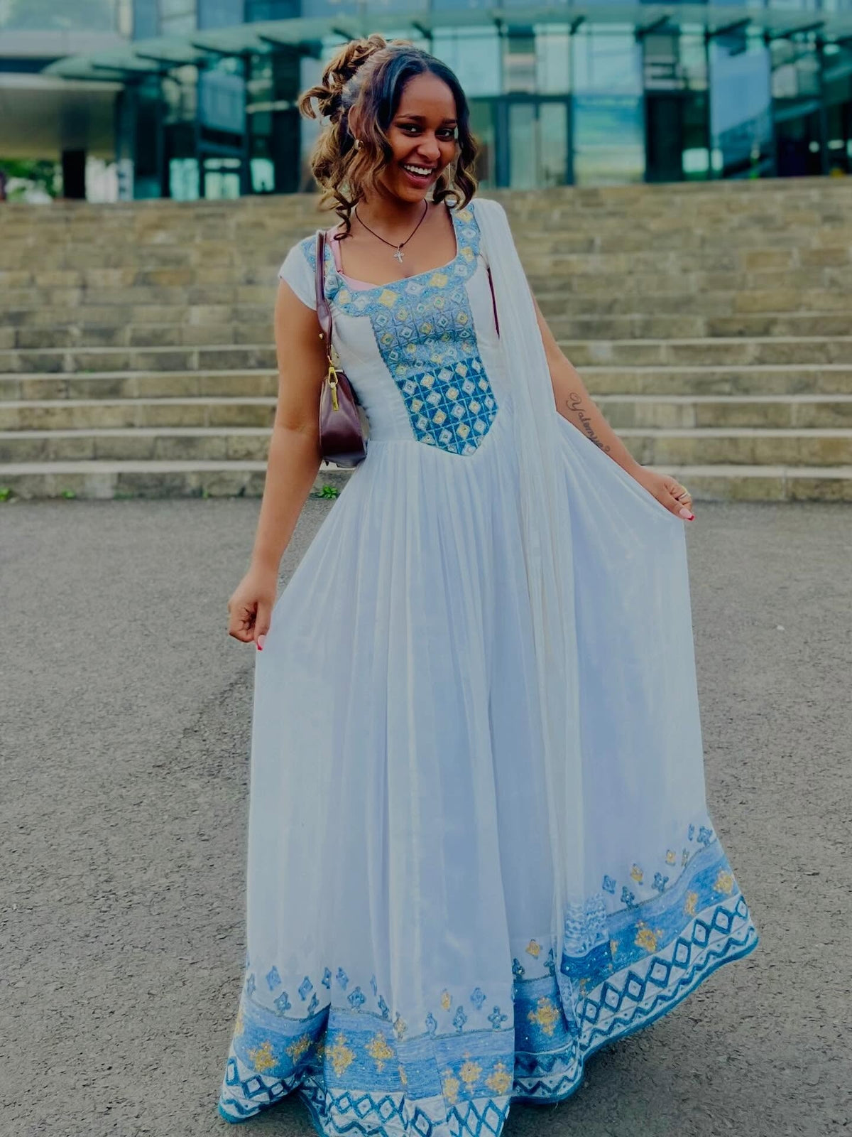 Woman in a long, light blue and white Habesha Kemis with floral patterns standing outdoors.