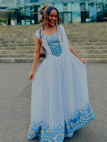 Woman in a long, light blue and white Habesha Kemis with floral patterns standing outdoors.