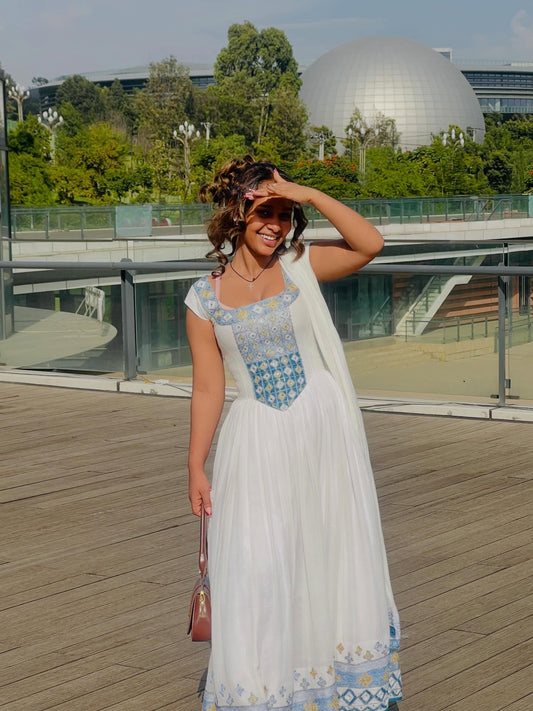 Woman in a white and blue Habesha Kemis standing on a wooden deck with greenery and a building in the background