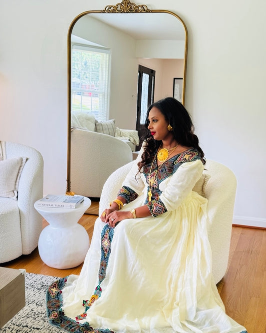 A woman sits in a cream boucle armchair in a bright living room, wearing a cream-colored traditional Habesha Kemis  with colorful embroidered details on the bodice and sleeves. A large floor mirror with an ornate gold frame stands behind her, reflecting the room interior.