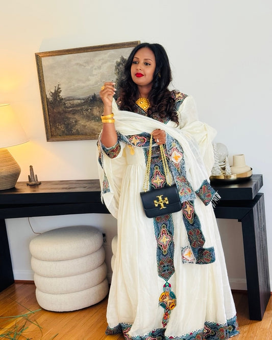 A woman models a traditional white embroidered Habesha Kemis  with colorful patterned borders, holding a black handbag with a gold cross logo. She is standing in a room with a console table, lamp, and framed painting in the background.
