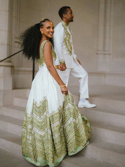 A woman in a white and sage green Green Habesha matching couple set traditional gown walks down stone steps while holding hands with a man in a matching white outfit with green embroidery.