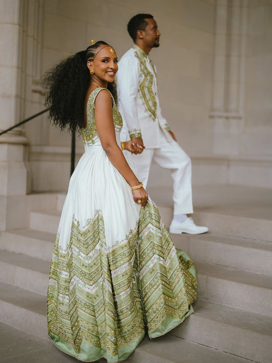 A woman in a white and sage green Green Habesha matching couple set traditional gown walks down stone steps while holding hands with a man in a matching white outfit with green embroidery.