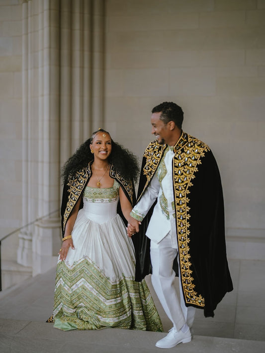 A couple in formal wedding Green Habesha matching couple set holding hands on stone steps. The bride wears a white strapless gown with green and beige patterned detailing and a black shawl with gold trim.