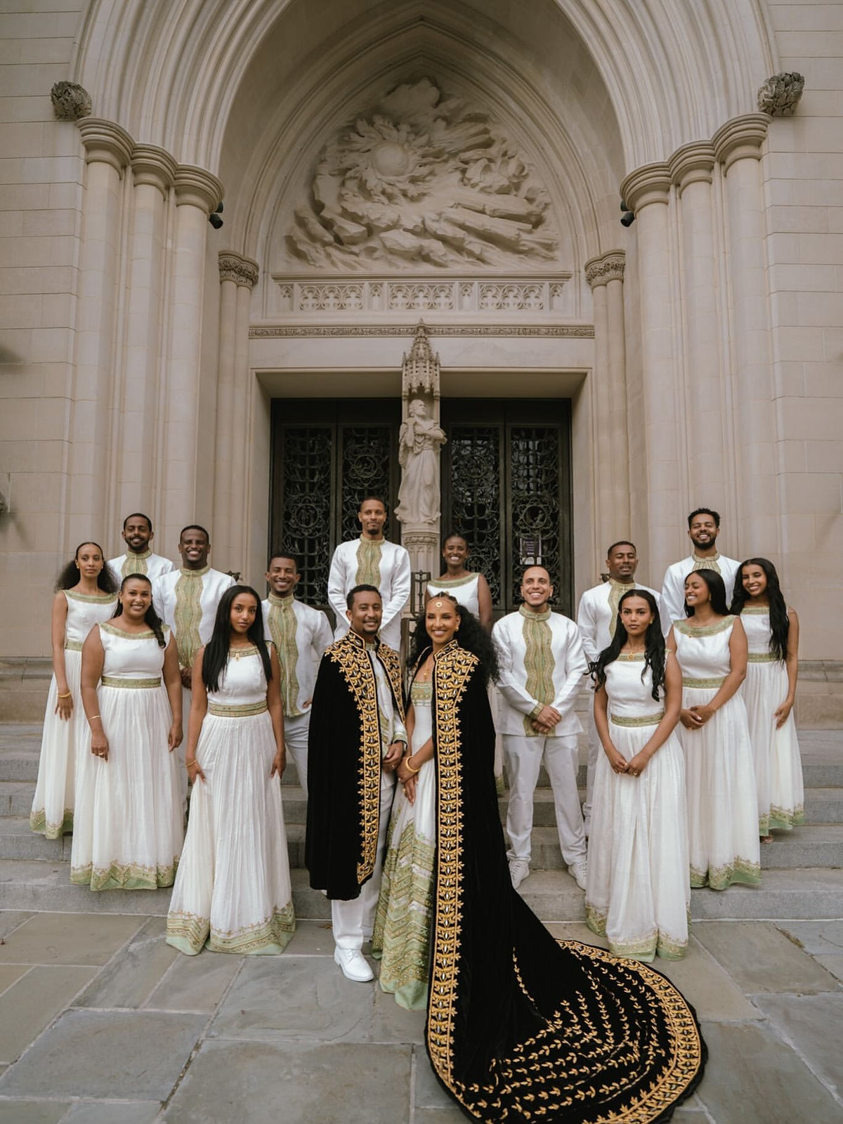 A wedding party poses in front of a grand stone cathedral doorway. The bride and groom stand in the center, wearing ornate black and gold ceremonial robes, while the brides Green Habesha matching couple set white sleeveless gowns with gold sashes and the groomsmen wear white suits with gold ties.