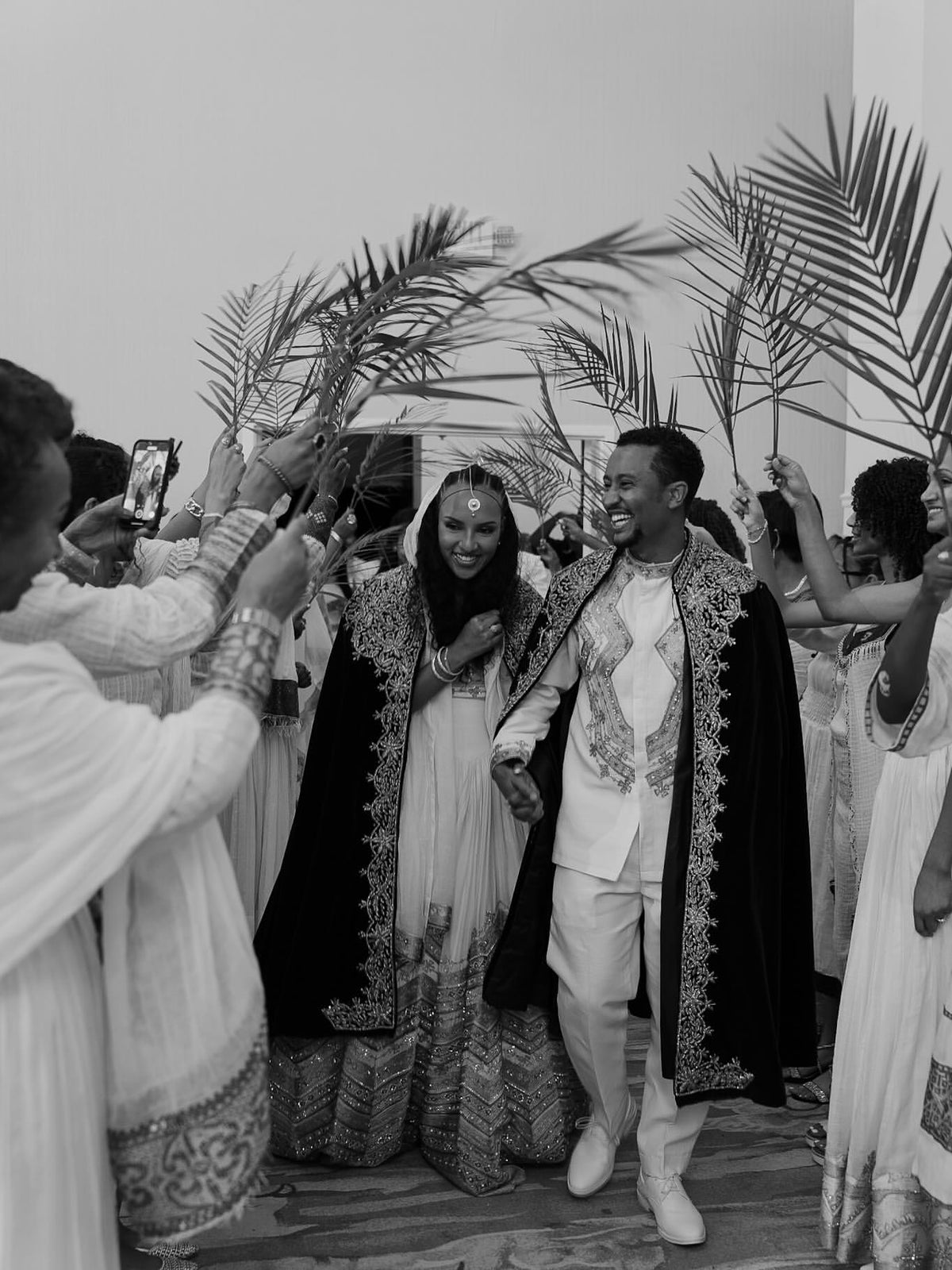 A black and white photograph of a couple walking Green Habesha matching couple set a aisle surrounded by guests holding palm leaves. The woman wears a light-colored traditional dress with a dark embroidered shawl, and the man wears a white traditional outfit with a dark embroidered shawl.